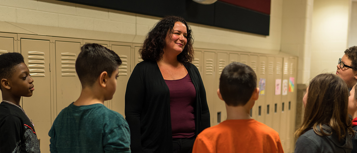 A woman standing in a hallway with lockers behind her and young students in front of her.