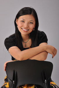 woman in a black shirt, leaning over the back of a chair, smiling at the camera