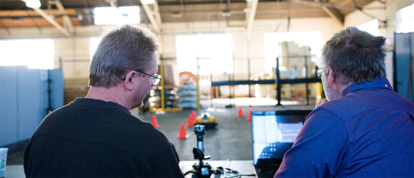 Two men operating a small remote controlled vehicle in a warehouse.