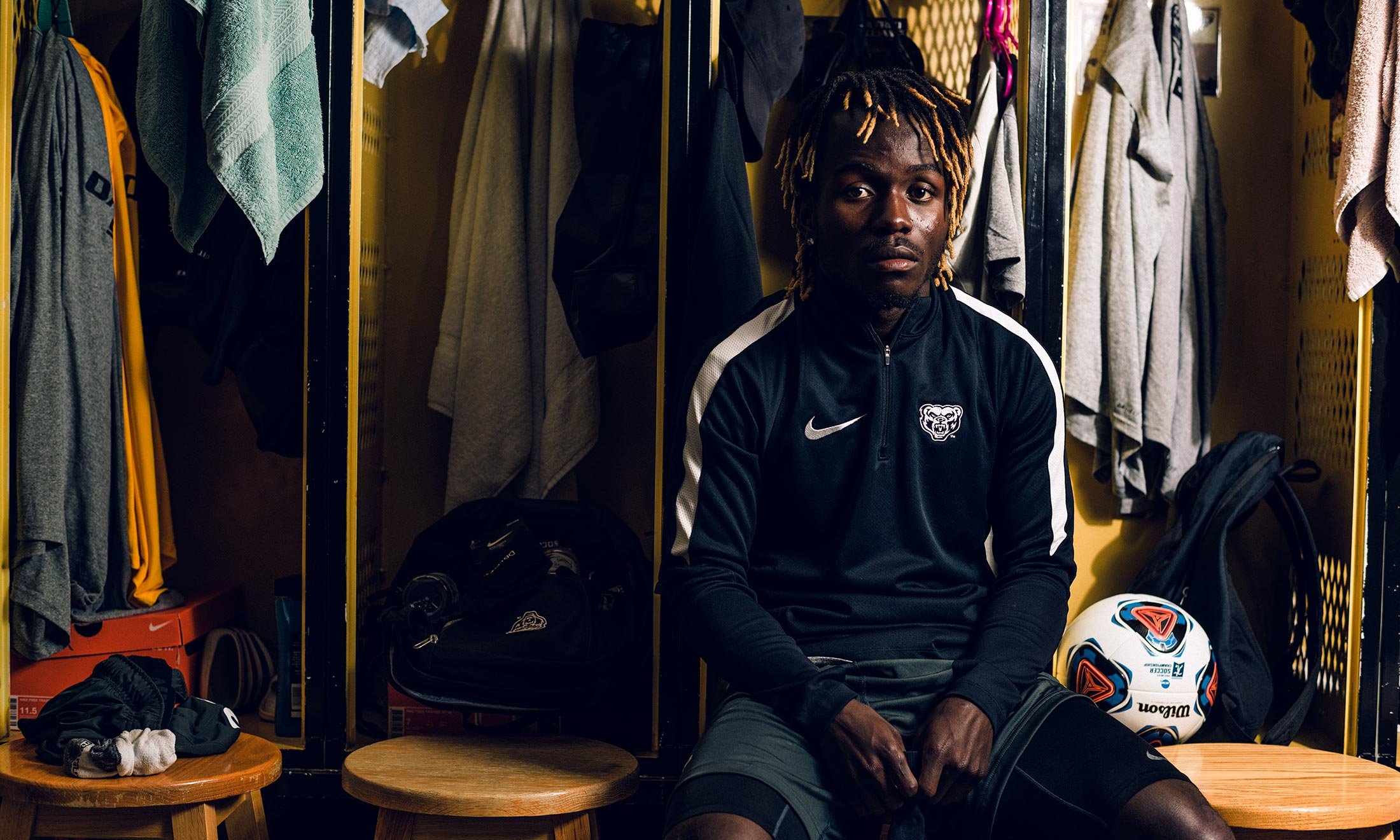 Oakland University soccer defender Wilfred Williams sits on a bench in the men's locker room with a soccer ball next to him with lockers and towels behind him