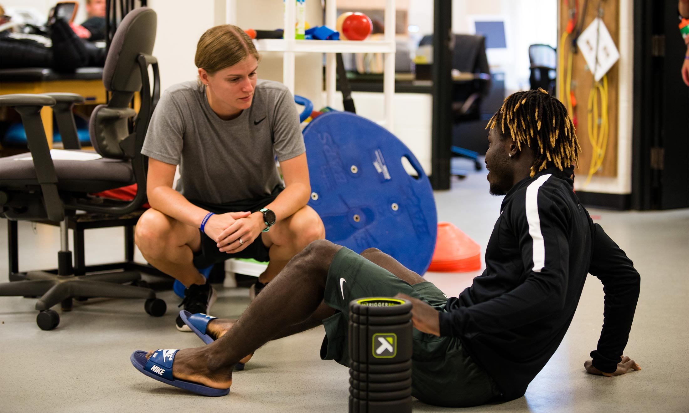 Oakland University soccer player Wilfred Williams sits on the floor to stretch while talking to an athletic trainer