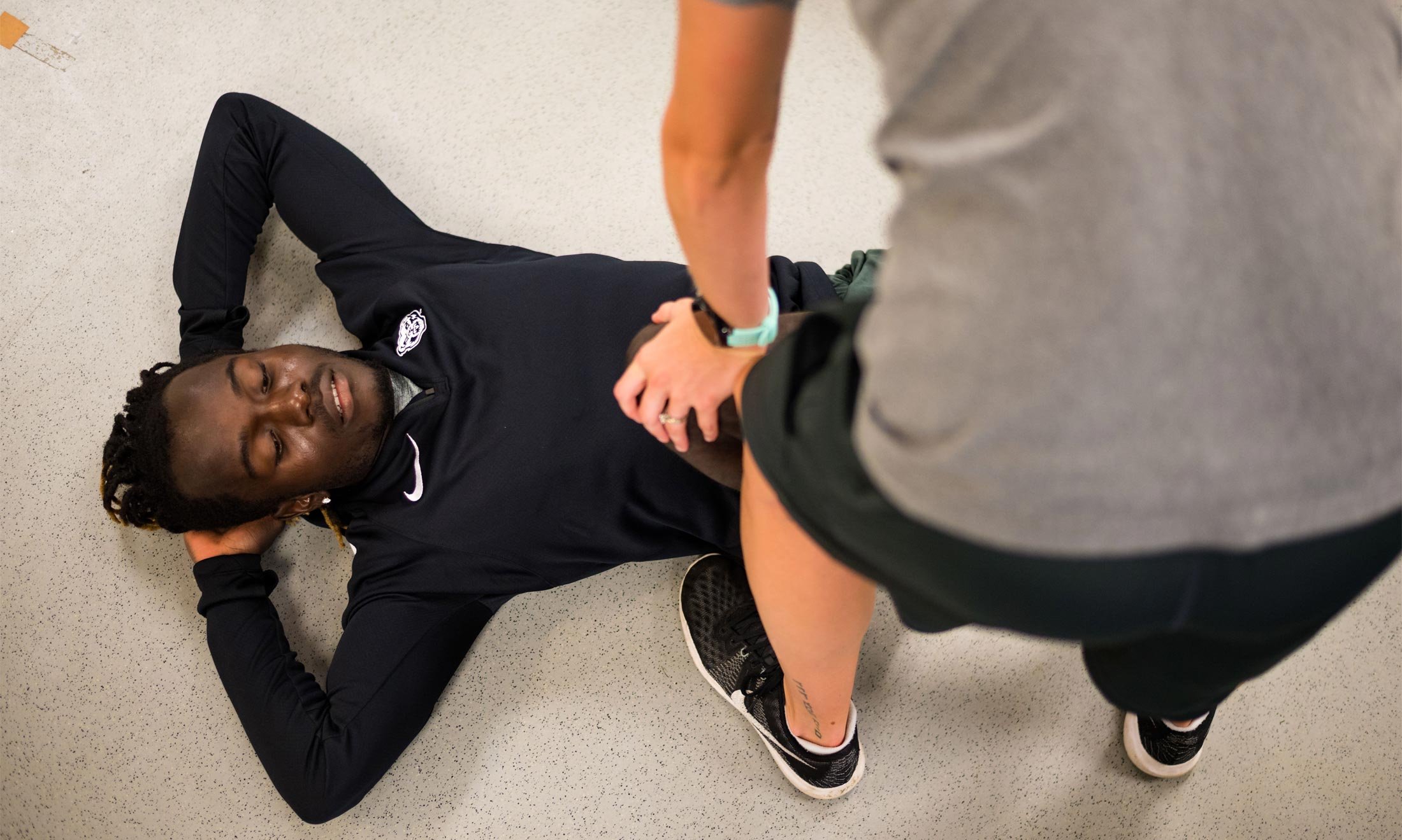 Oakland University soccer player Wilfred Williams lays on a white floor while a trainer helps stretch his legs