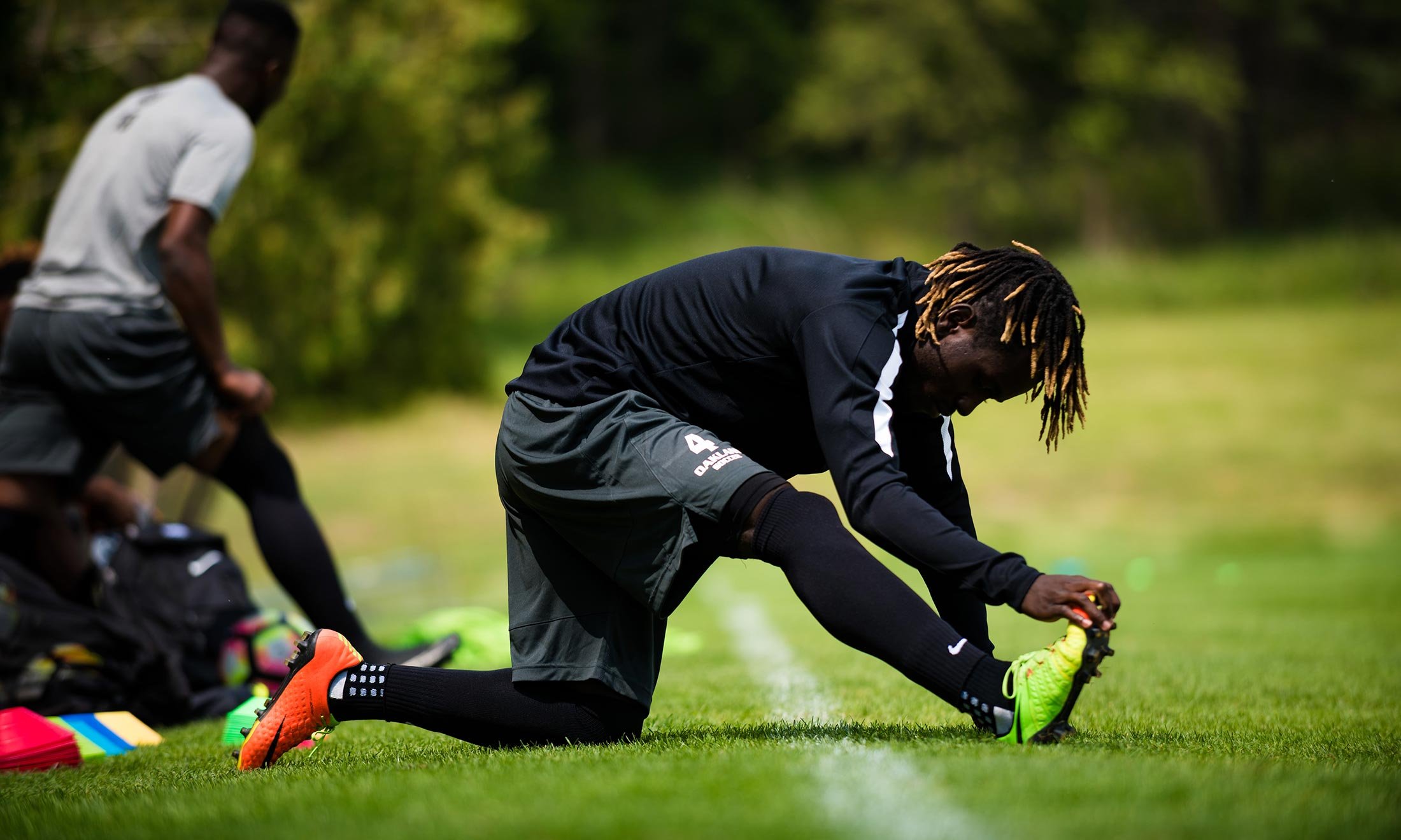 Soccer player Wilfred Williams stretches his right leg on the grass of the Oakland University soccer practice field while other teammates practice behind him