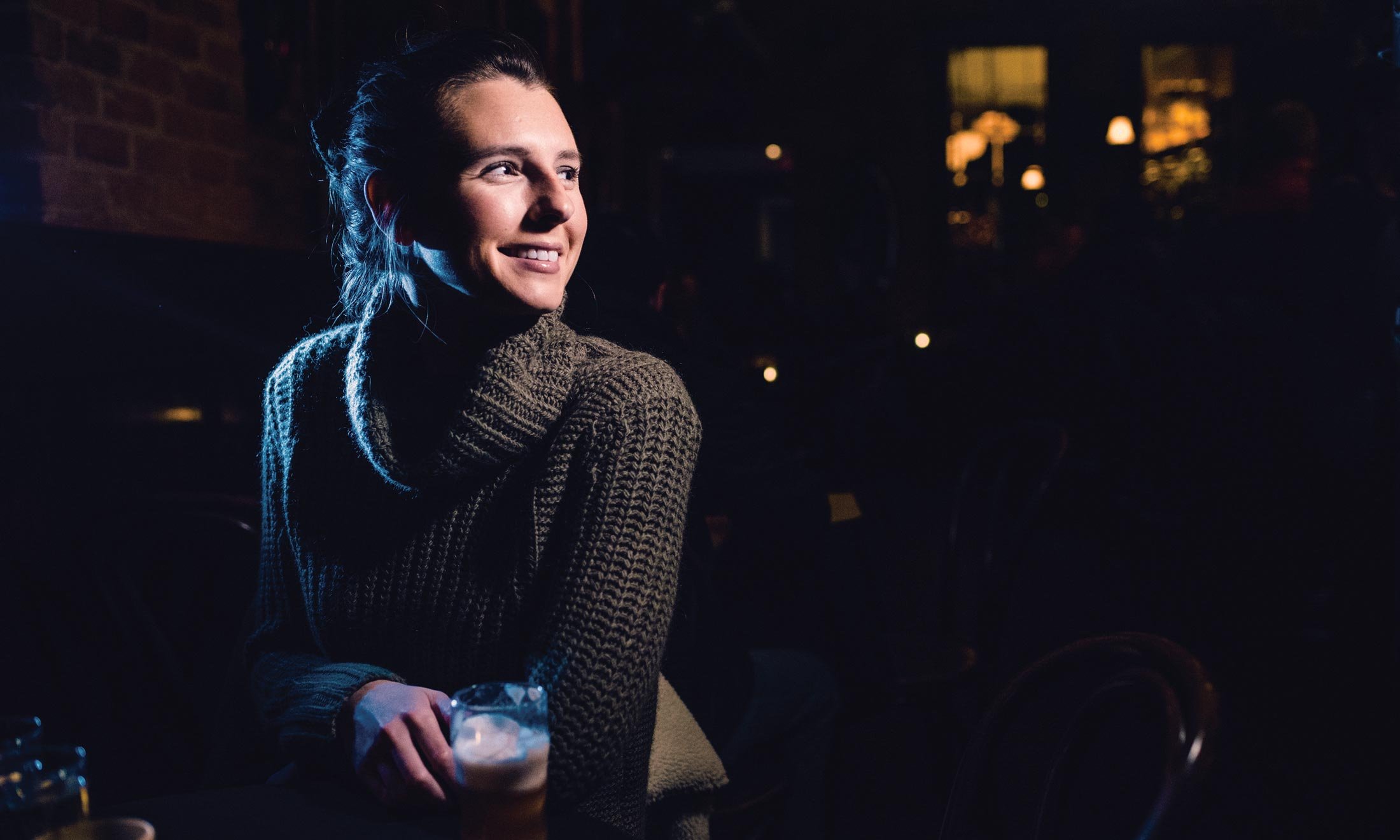 Oakland University alumna Hillary Sawchuck, creator of 'A Drink With,' has a drink at Sugar House in Detroit, with a dark and blurry view of the pub interior in the background.