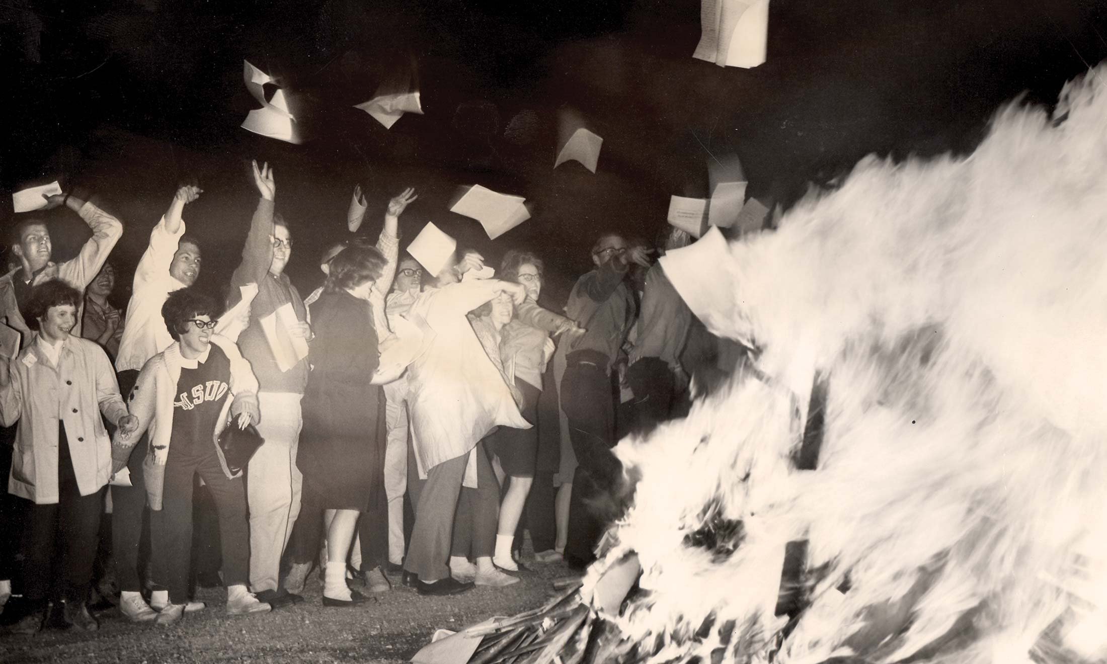 Black and white photo of Oakland University students tossing their blue book exam booklets into a large bonfire.