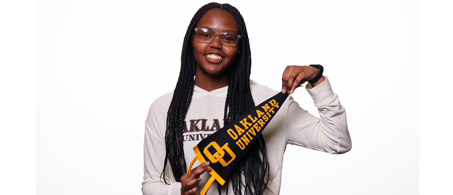 Young woman smiles at the camera. She is wearing and Oakland University shirt and is holding an O U pennant.