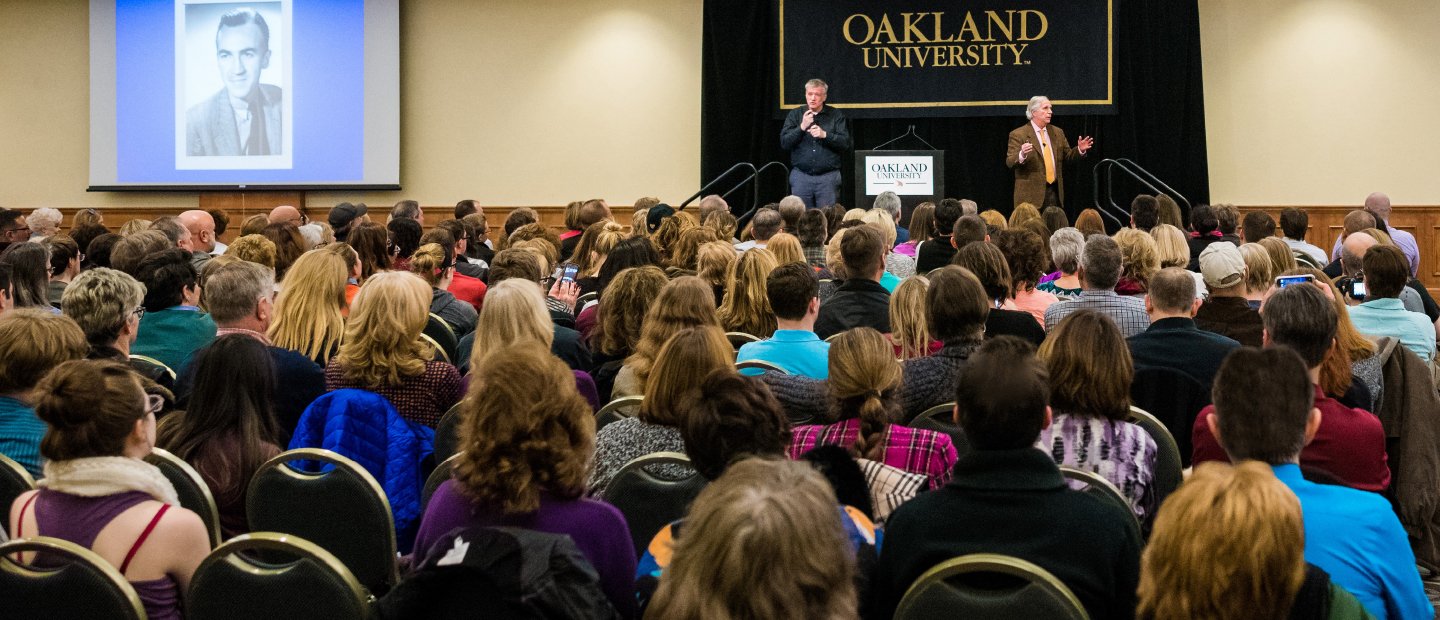 two men standing in front of a black Oakland University banner, addressing a seated audience
