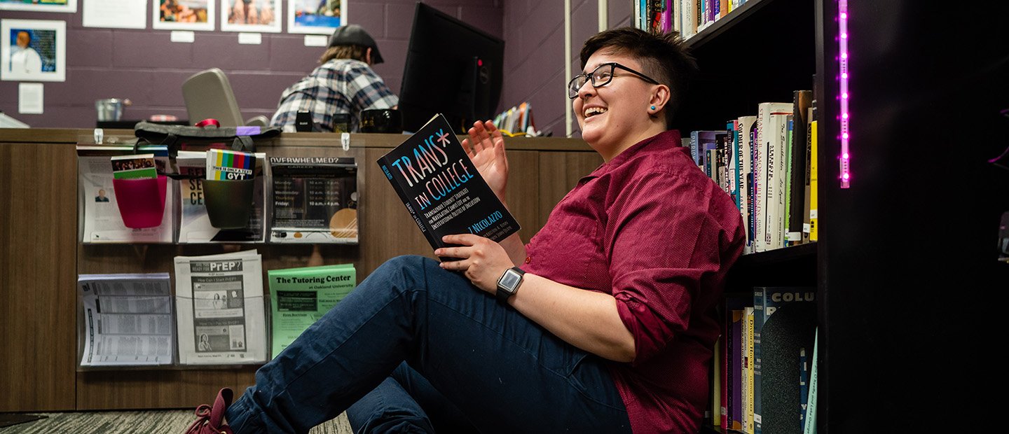 young adult seated in front of a book shelf reading a book called "Trans in College"
