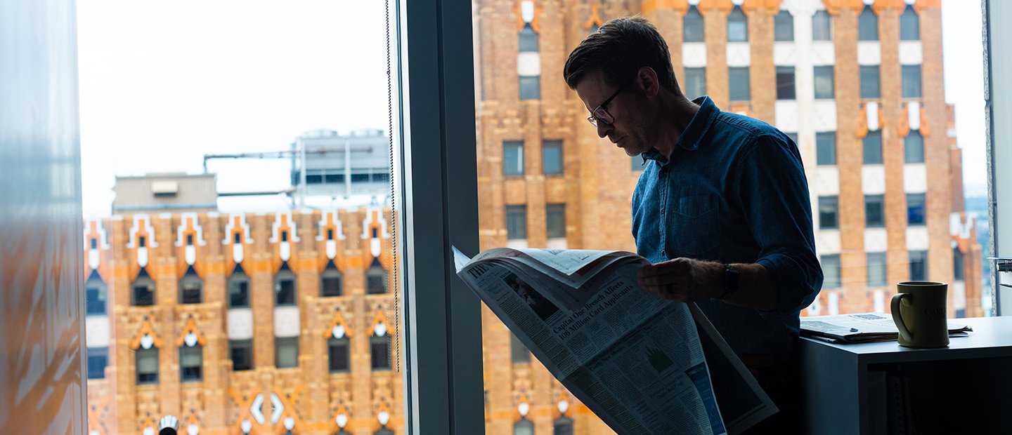 man reading a newspaper, standing in front of a window