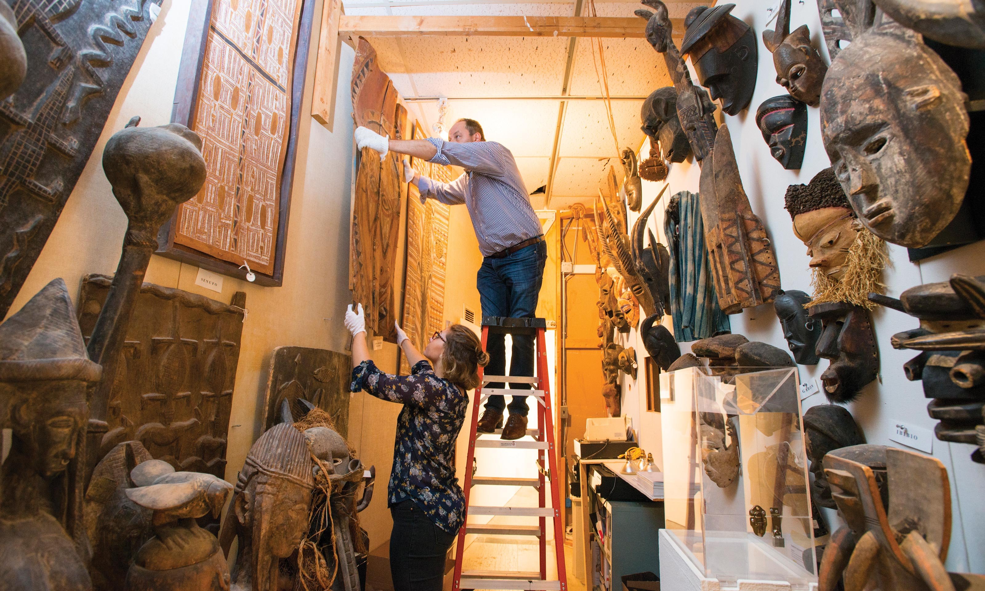 O U Art Gallery director Dick Goody stands on a ladder to hang some African art with the help of a student. African masks decorate the walls. 