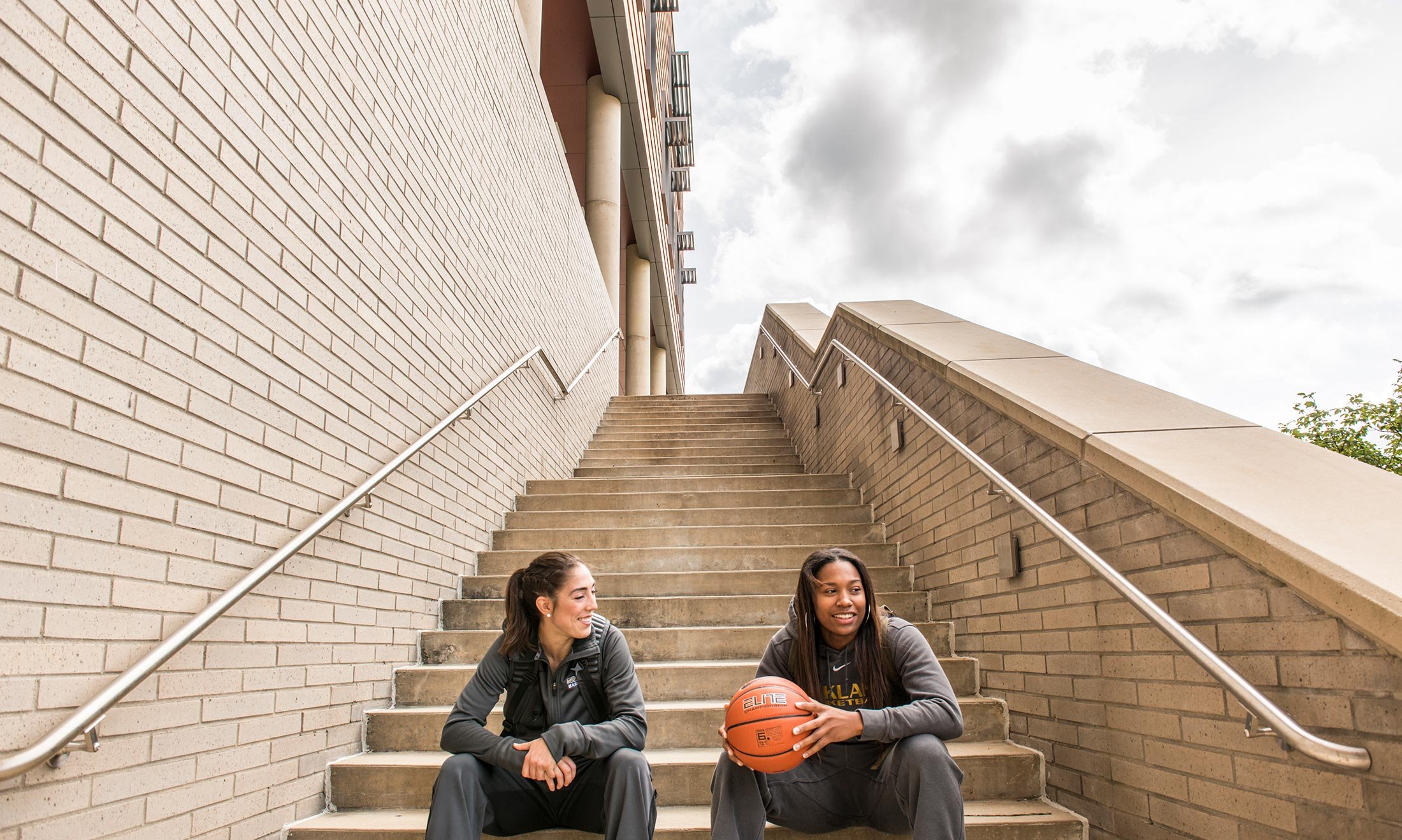 Oakland University women's basketball player Taylor Gleason sits on the steps of the Human Health building and talks with a friend holding a basketball 