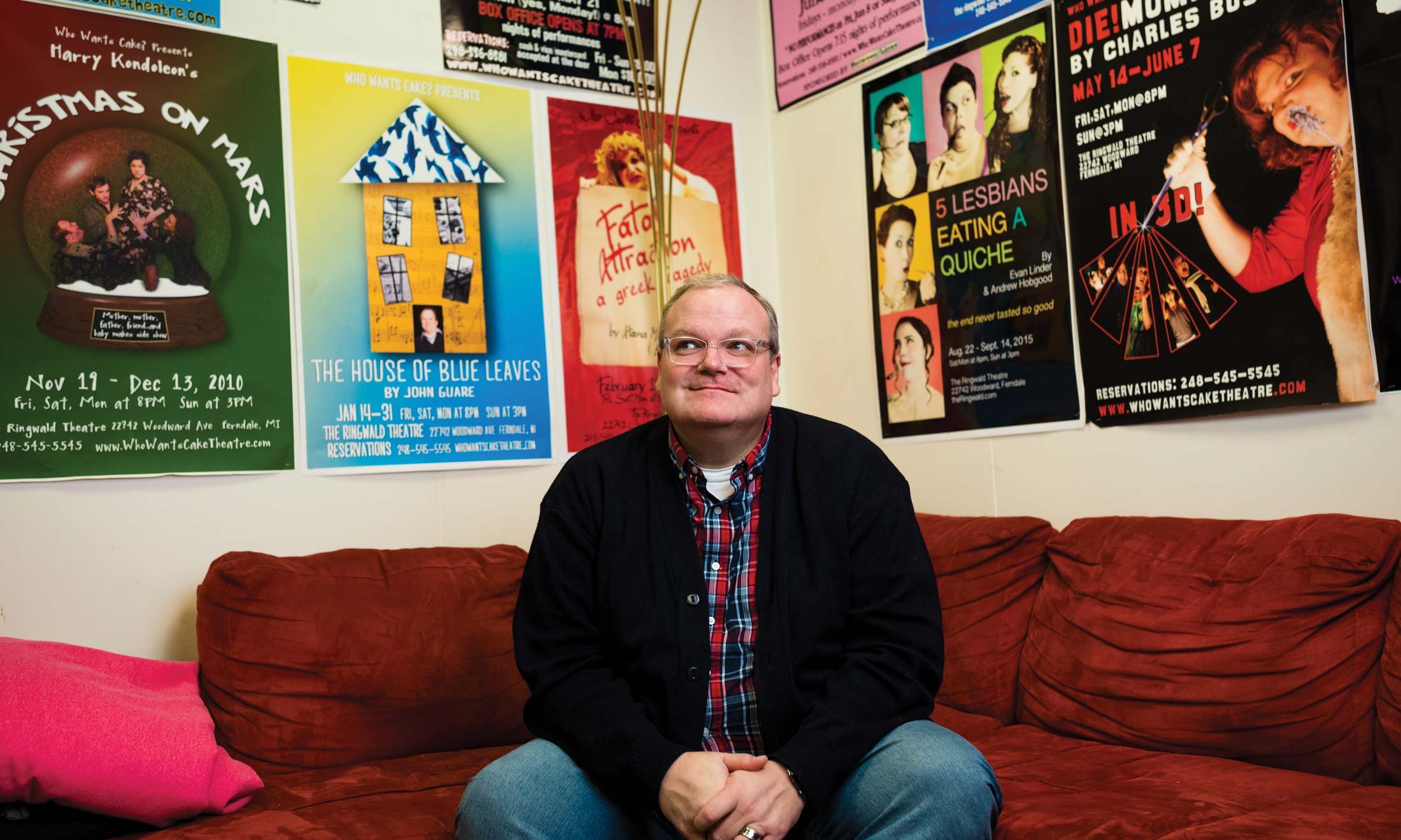 Backstage at The Ringwald Theatre in Ferndale, Michigan, Joe Bailey sits on a red couch in his office. Many posters from his shows line the walls. Wearing a black jacket, jeans and a flannel shirt.