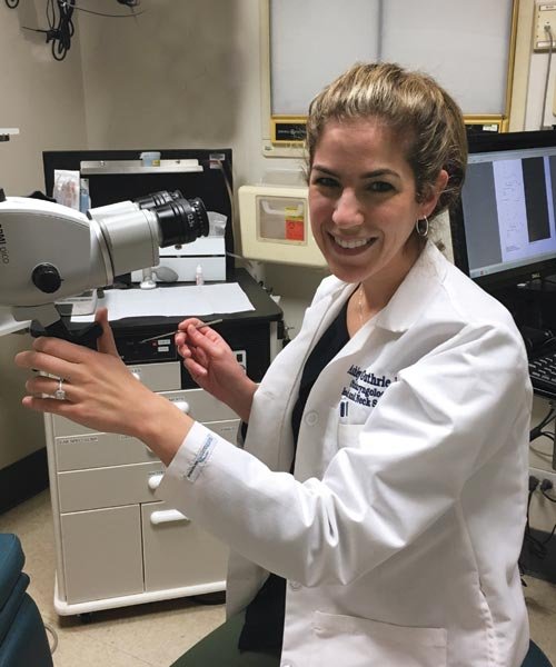 O U W B graduate Dr. Ashley J. Guthrie, standing in a hospital room next to a microscope. Wearing her white lab coat, smiling. 