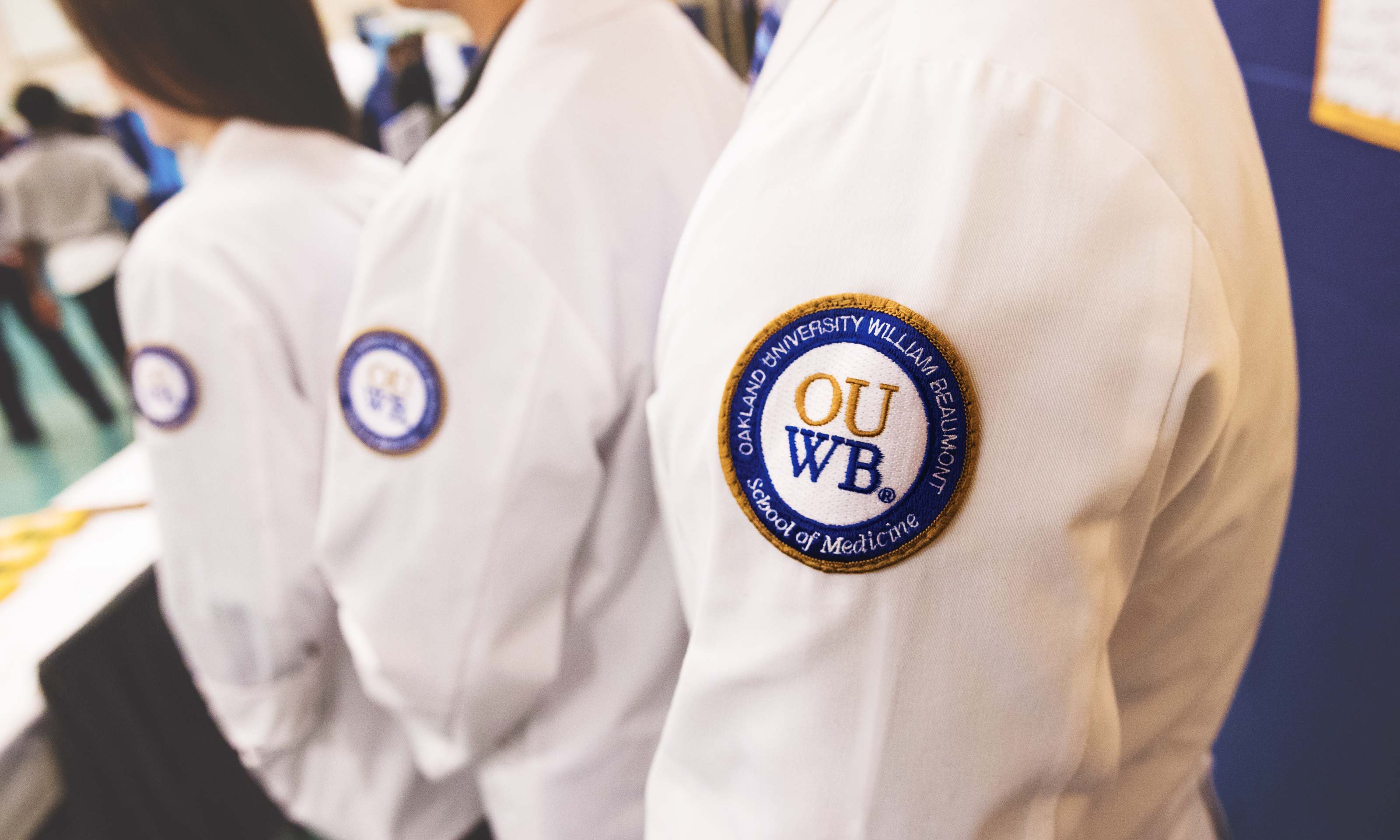 O U W B students in white coats. The O U W B logo crest is in the front of the photo, on the white coats.
