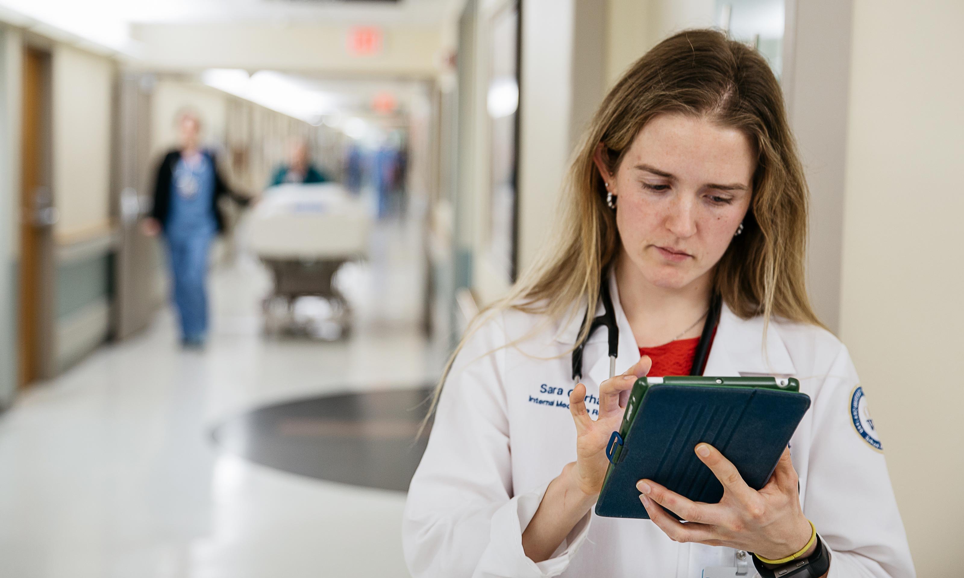 O U W B graduate Dr. Sara Gerhardt serving her residency at Beaumont Hospital in Royal Oak, Michigan. Standing in the hospital hallway, looking at an i pad, wearing a white coat.