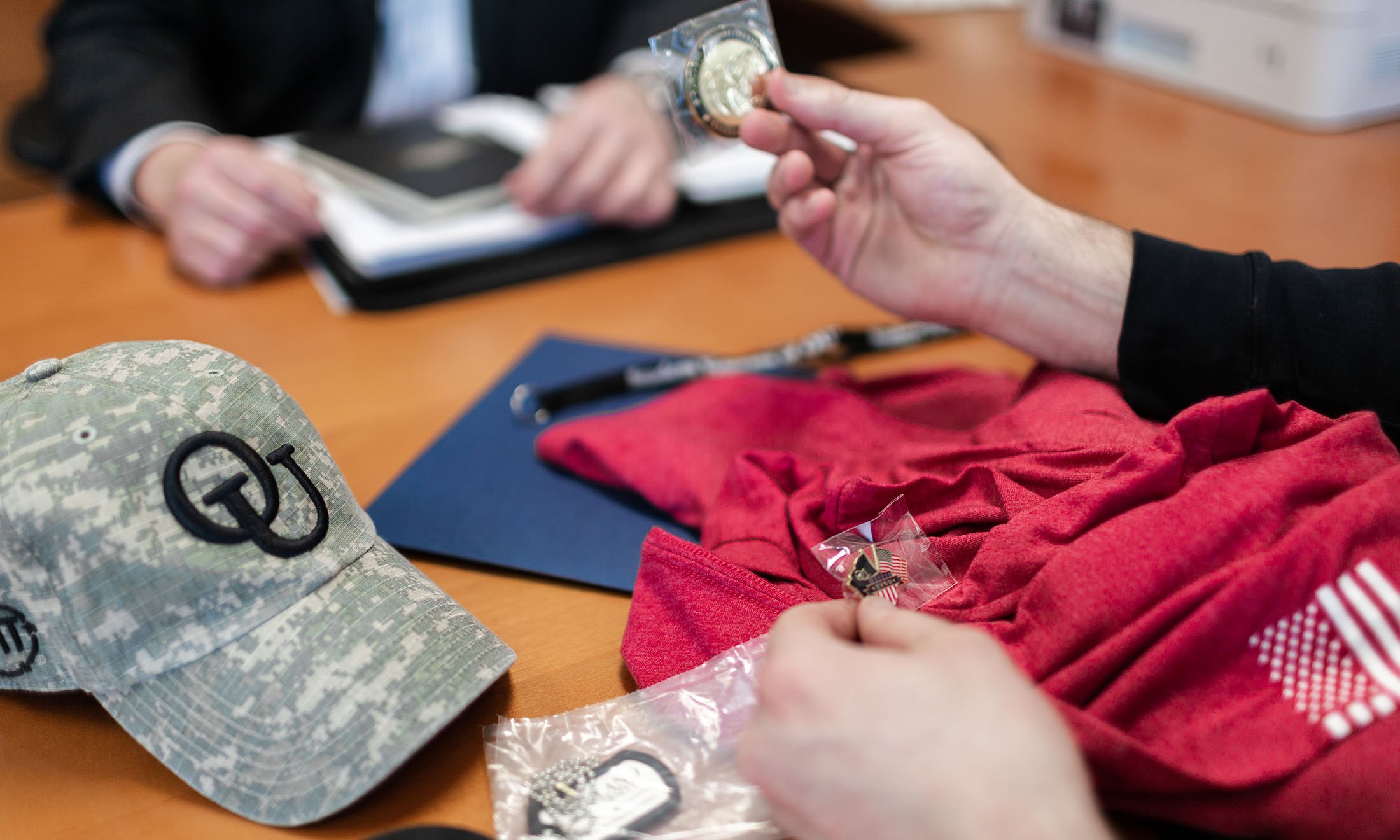 Hands holding OU Veteran Support Services pin and coin, over desk with red t-shirt, camouflage OU hat, and dog tags.