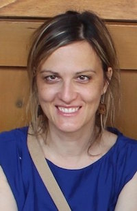 woman in a blue shirt in front of a wood paneled wall, smiling at the camera