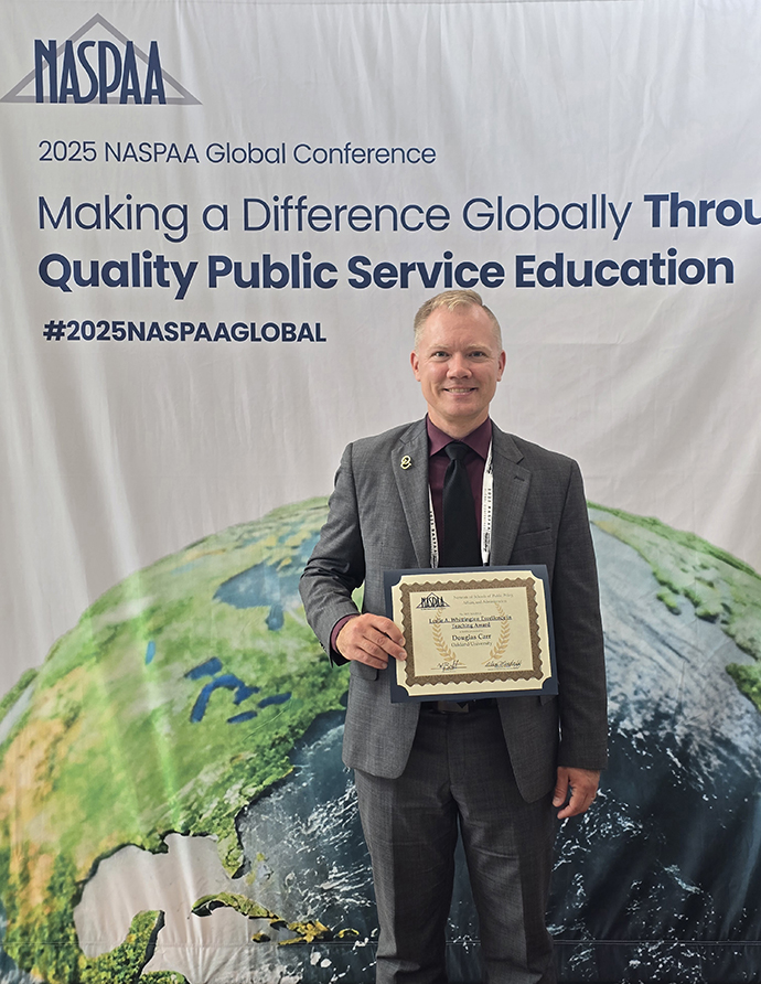 Doug Carr holds up his certificate in front of the NASPAA banner.