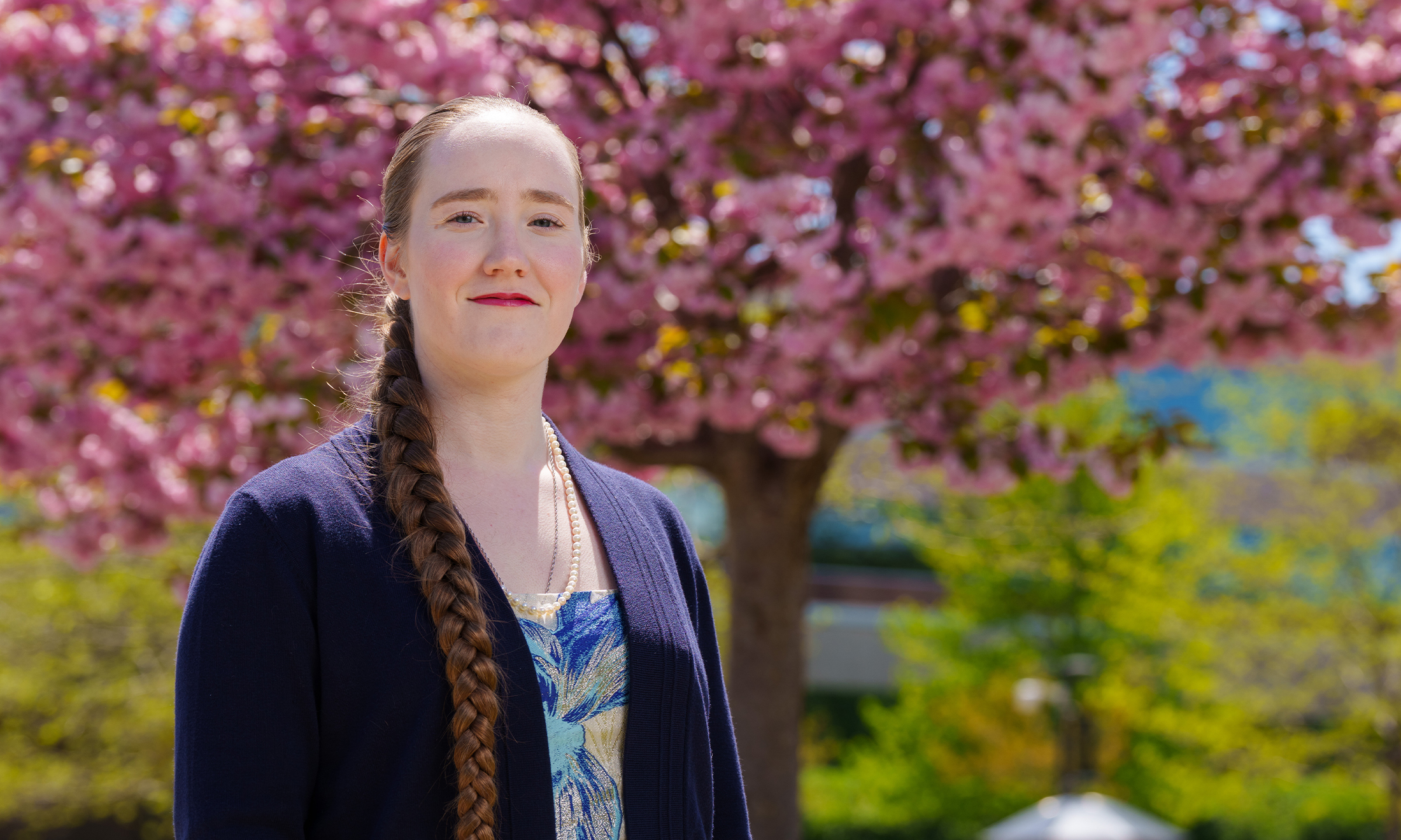 A woman stands facing the camera. There is a flowering tree in the background.