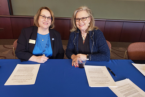 Two women sitting at a table with papers. They smile at the camera.