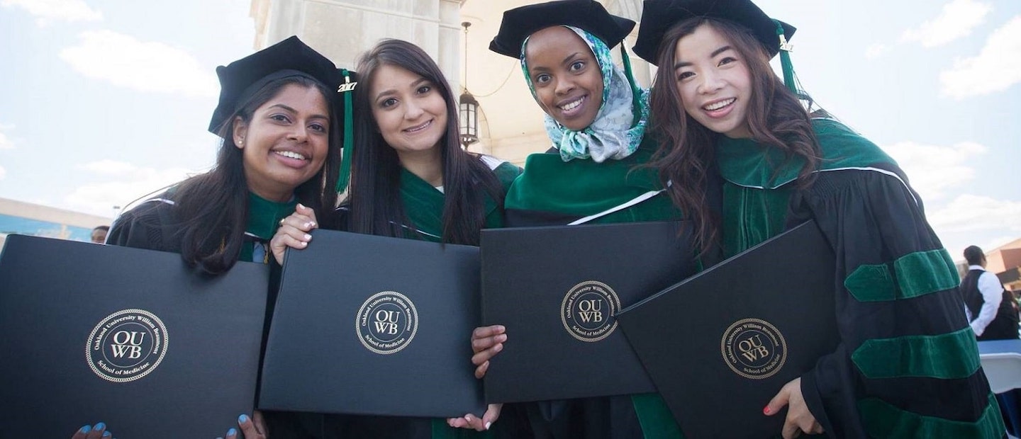 Four new OUWB graduates stand before the Elliot Tower wearing caps and gowns and holding their diplomas.