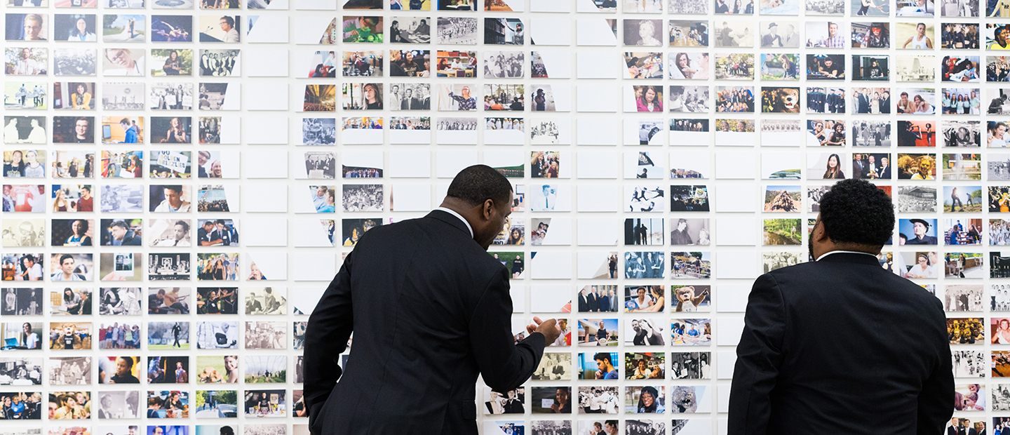 Two people view a wall of photographs with the O U logo on it.