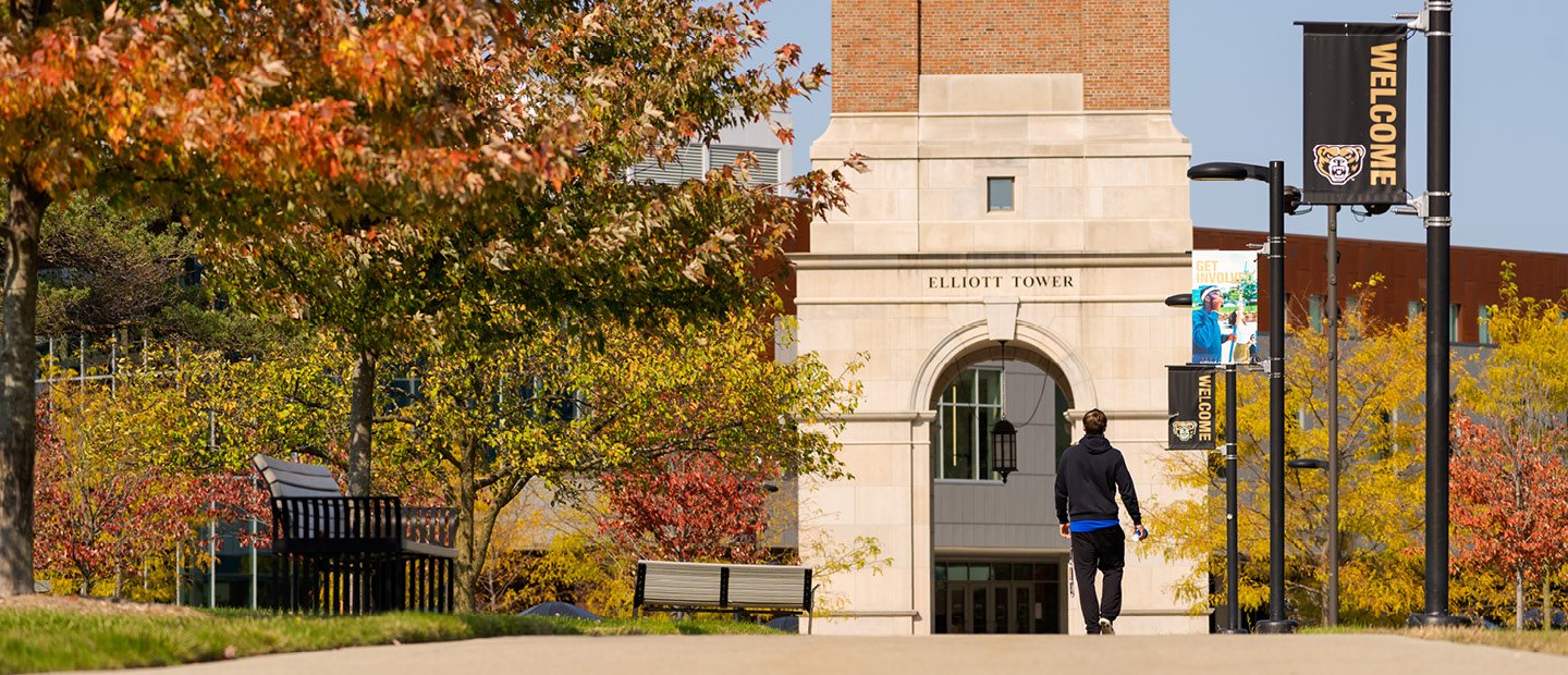 A person walking on a sidewalk towards Elliott Tower on Oakland University's campus.